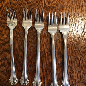 Puede incluir: Cinco tenedores de plata con mangos ornamentados, dispuestos en fila sobre una superficie de madera.