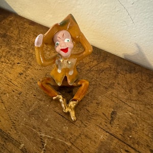 May include: A ceramic figurine of a jester with a green and brown hat, wearing a brown and white patterned outfit, with gold accents on the shoes and hat. The jester has a painted face with green eyes, a red mouth, and a pink nose.