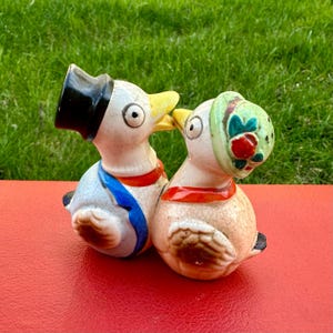 Puede incluir: Un par de saleros y pimenteros de cerámica con forma de pájaros. El pájaro de la izquierda lleva un sombrero de copa negro y un chaleco azul y blanco. El pájaro de la derecha lleva un sombrero verde con flores rojas. Los pájaros se besan.