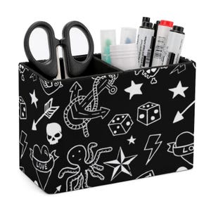 May include: Black desk organizer with a white tattoo-style pattern featuring anchors, stars, dice, and skulls. The organizer holds scissors, pens, and other office supplies. A stylish and functional desk accessory.
