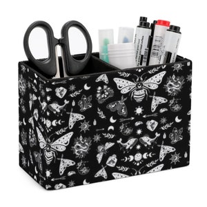May include: Black desk organiser with a white pattern of moths, moons, stars, and other celestial designs. The organiser has three compartments and is filled with scissors, pens, and markers.