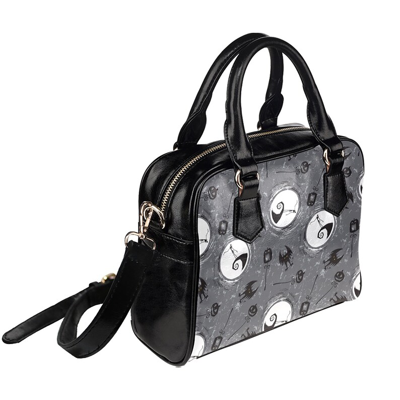 K&ouml;nnte beinhalten: Schwarze Handtasche mit Schultergurt. Die Tasche hat ein grau-wei&szlig;es Muster mit einem sich wiederholenden Bild von Jack Skellington aus Nightmare Before Christmas.