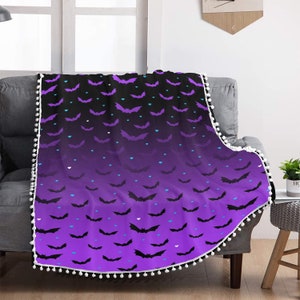 Può includere: Coperta in pile viola e nera con un motivo a forma di pipistrello e cuore. La coperta ha un bordo di pon pon bianchi.