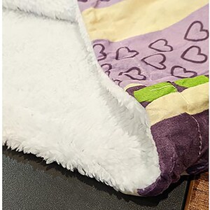 Może przedstawiać: Miękki, biały koc z polaru z purpurowo-ż&oacute;łtym paskowanym wzorem i serduszkami. Koc jest złożony na p&oacute;ł, a strona z polaru jest zwr&oacute;cona w stronę aparatu.