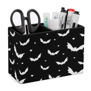 May include: Black desk organizer with a bat pattern and white hearts. The organizer has three compartments and is holding scissors, markers, and other office supplies.