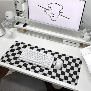May include: A white desk with a black and white chequered mouse mat, a white keyboard, a white computer monitor, and a white mouse. The monitor has a cartoon drawing of a bear on it.