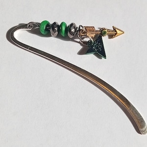 May include: A silver metal bookmark with a curved design. It features green and silver beads, a gold-colored arrow charm with a green gem, and a dark green star charm. The bookmark is on a white background.