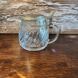May include: A clear glass beer mug featuring a swirling, textured pattern on the exterior body. The mug includes a sturdy glass handle and a hinged, circular metal lid with a decorative thumb lever attached to the top rim of the vessel.