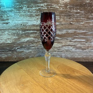 May include: A tall, slender champagne flute featuring a deep red glass bowl with a clear, cut-glass diamond lattice pattern and a starburst motif near the rim. The stem and circular base are made of clear, smooth glass, providing a stable foundation.