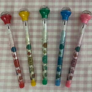 May include: Five colorful pencils with clear barrels and decorative animal and fruit designs. Each pencil has a different colored top: red, yellow, green, blue, and pink. The pencils are arranged on a pink and white checkered surface.