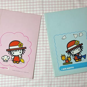 May include: Two notebooks with cartoon illustrations. One is pink with a character in a red dress and hat, a dog, and the text "I AM HATTY". The other is light blue with a similar character playing a trumpet and the same text.