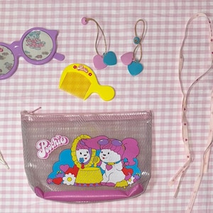 May include: A collection of vintage children's accessories, including a purple toy pair of sunglasses, a yellow comb, pink and blue earrings, a clear pouch with a cartoon graphic, a ribbon, and a small card that says "Hair Fun".