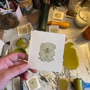 May include: A square, off-white print featuring a green image of a Green Man face with a beard and leafy hair. The print is held in a hand, with other prints and art supplies visible in the background.