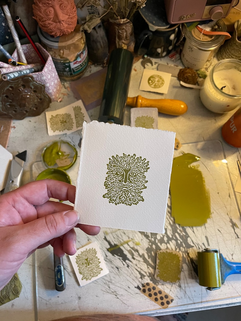 May include: A square, off-white print featuring a green image of a Green Man face with a beard and leafy hair. The print is held in a hand, with other prints and art supplies visible in the background.