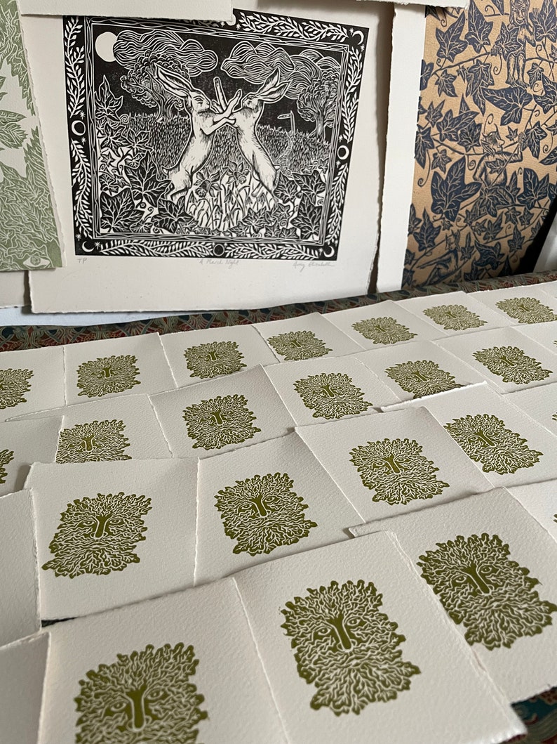 May include: A collection of art prints featuring a Green Man design and a black and white image of two hares. The Green Man prints are in olive green ink on white paper. The hare print is framed with leaves and moons.
