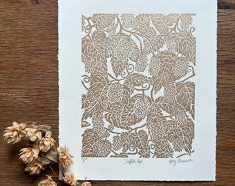 May include: A tan and white print featuring a detailed botanical illustration of hops. The artwork, titled "Suffolk Hops" by Amy Elizabeth, showcases a repeating pattern of leaves and cones. The print is on textured paper, with dried hops in the foreground.