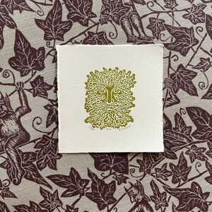 May include: A square, off-white print featuring a green image of the Green Man with a face surrounded by leaves and branches. The print is on textured paper. The background is a fabric with a repeating pattern of dark brown ivy leaves and figures.