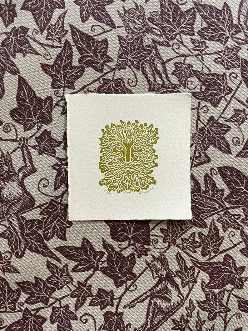 May include: A square, off-white print featuring a green image of the Green Man with a face surrounded by leaves and branches. The print is on textured paper. The background is a fabric with a repeating pattern of dark brown ivy leaves and figures.
