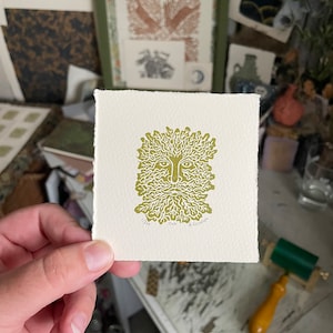 May include: A square print featuring a stylized green face with a beard and leafy hair. The print is on off-white paper and is held in a hand. The print is labeled "Oak" and "1/300". Other prints and art supplies are in the background.