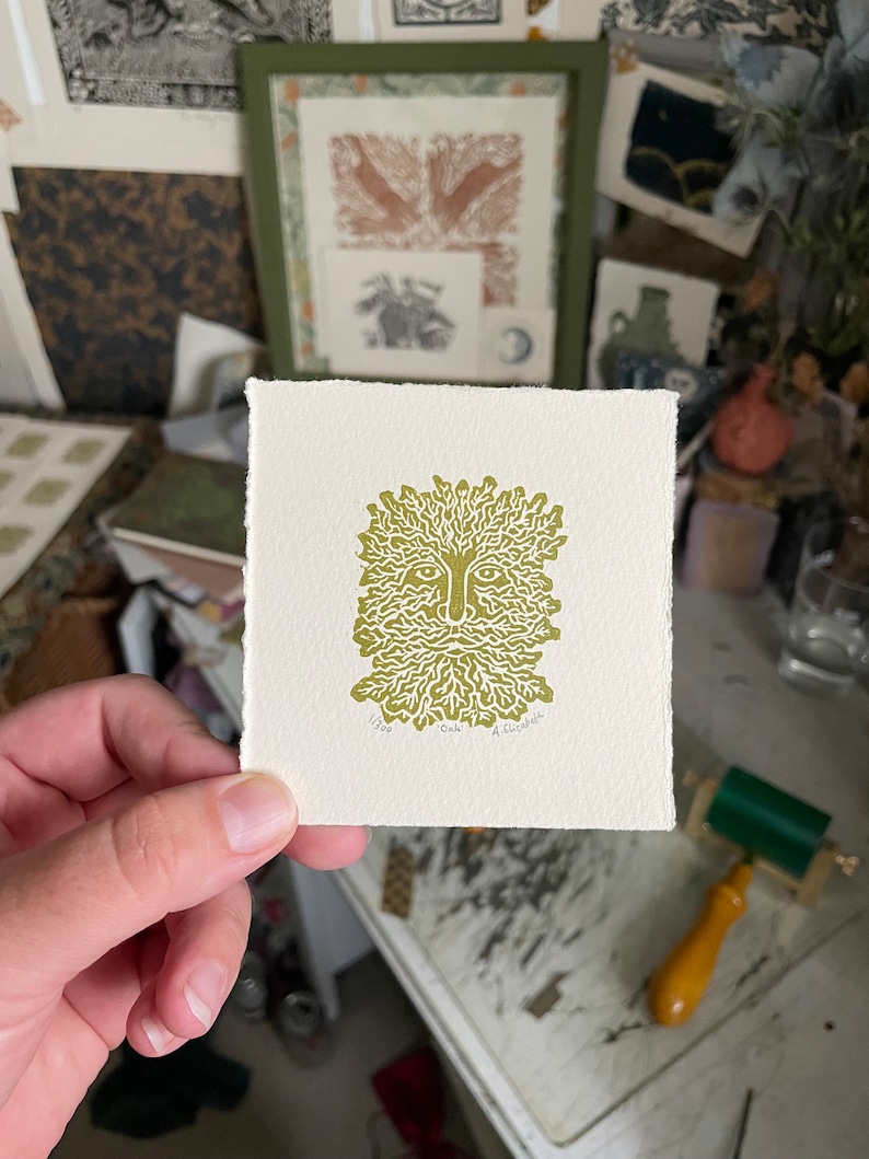 May include: A square print featuring a stylized green face with a beard and leafy hair. The print is on off-white paper and is held in a hand. The print is labeled "Oak" and "1/300". Other prints and art supplies are in the background.