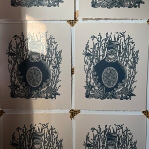 Puede incluir: Varias impresiones de un dise&ntilde;o de botella azul oscuro, rodeado de algas marinas, estrellas de mar y conchas. Las impresiones est&aacute;n sobre papel blanquecino y fijadas con cinta adhesiva decorativa. La obra tiene un tema n&aacute;utico.