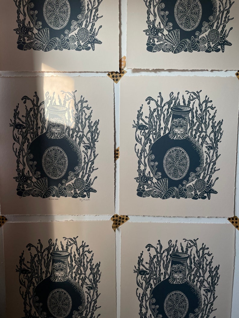 Puede incluir: Varias impresiones de un dise&ntilde;o de botella azul oscuro, rodeado de algas marinas, estrellas de mar y conchas. Las impresiones est&aacute;n sobre papel blanquecino y fijadas con cinta adhesiva decorativa. La obra tiene un tema n&aacute;utico.