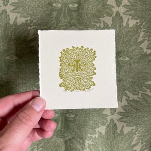 May include: A square print featuring a Green Man design in olive green ink on textured off-white paper. The Green Man has a face with a beard and leafy hair. The print is held against a green fabric with a repeating Green Man face pattern.