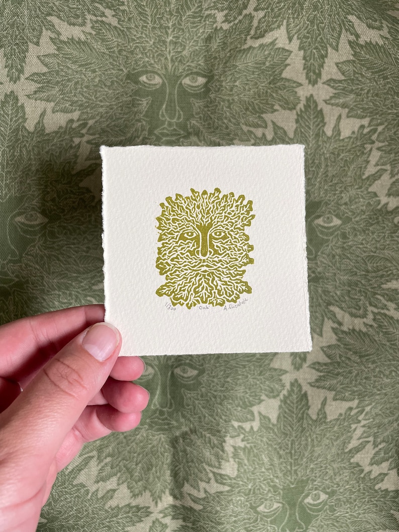 May include: A square print featuring a Green Man design in olive green ink on textured off-white paper. The Green Man has a face with a beard and leafy hair. The print is held against a green fabric with a repeating Green Man face pattern.