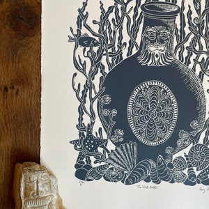 Puede incluir: Un grabado en lin&oacute;leo azul oscuro y blanco titulado "The Witch Bottle" que representa una botella con una cara, rodeada de algas, peces, estrellas de mar y conchas. La impresi&oacute;n est&aacute; sobre papel texturizado. Una cara de piedra tallada est&aacute; en primer plano.