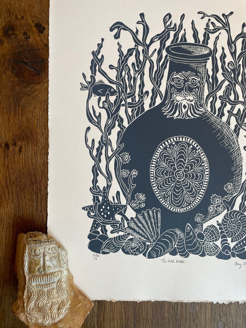 Puede incluir: Un grabado en lin&oacute;leo azul oscuro y blanco titulado "The Witch Bottle" que representa una botella con una cara, rodeada de algas, peces, estrellas de mar y conchas. La impresi&oacute;n est&aacute; sobre papel texturizado. Una cara de piedra tallada est&aacute; en primer plano.