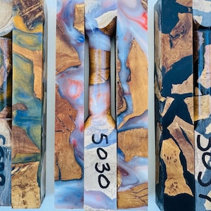 May include: Three rectangular blocks of wood and resin composite, each featuring a central turned cylindrical section. The blocks are labeled with the numbers 5039, 5030, and 5057. The materials display a mix of natural wood grain and swirls of orange, blue, white, red, and black resin.