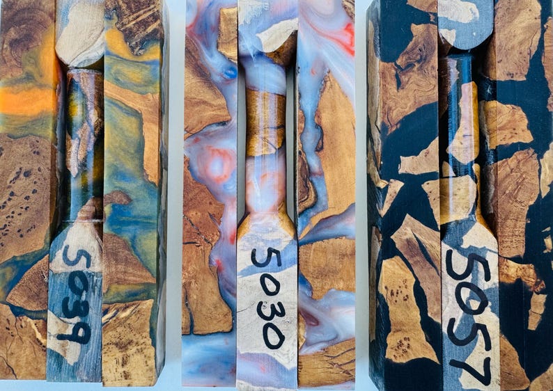 May include: Three rectangular blocks of wood and resin composite, each featuring a central turned cylindrical section. The blocks are labeled with the numbers 5039, 5030, and 5057. The materials display a mix of natural wood grain and swirls of orange, blue, white, red, and black resin.