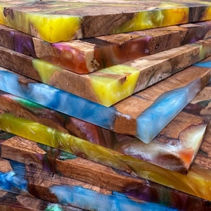 May include: A stack of handcrafted wooden boards with colorful epoxy resin accents. The boards feature a mix of natural wood grain and vibrant resin in shades of yellow, blue, purple, and orange. The boards are likely for home decor or woodworking projects.