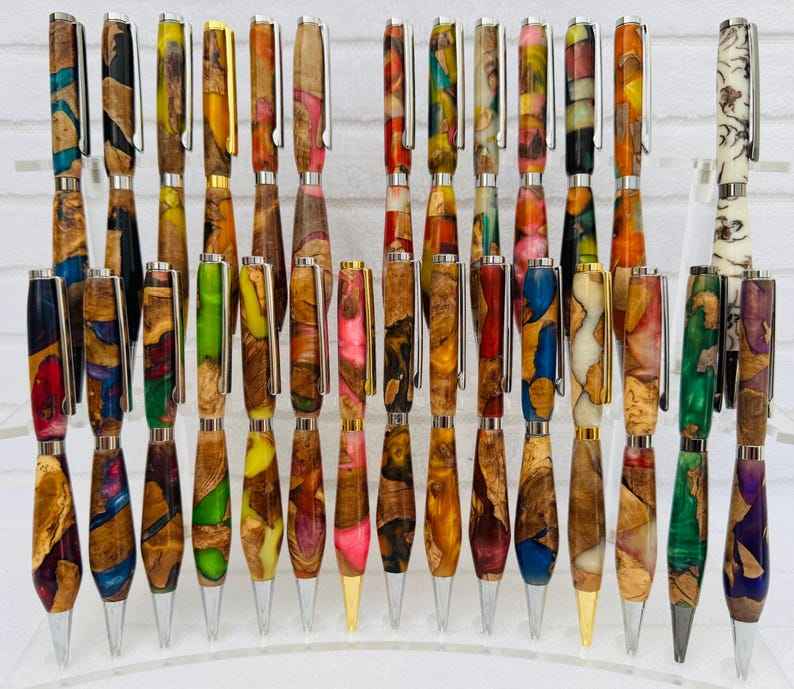 May include: A collection of handcrafted pens arranged in rows, featuring barrels made from a combination of natural wood and colorful resin. Each pen includes a metal clip and a pointed tip, showcasing various swirling patterns and vibrant color combinations.