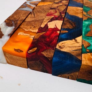 May include: Five rectangular blocks of wood combined with colored resin. The blocks feature natural wood grain patterns interspersed with swirls of white, orange, red, blue, and green resin, creating a distinct contrast between the organic wood and the vibrant, semi-translucent synthetic material.