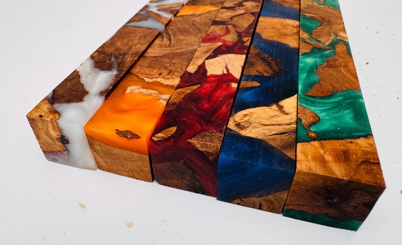 May include: Five rectangular blocks of wood combined with colored resin. The blocks feature natural wood grain patterns interspersed with swirls of white, orange, red, blue, and green resin, creating a distinct contrast between the organic wood and the vibrant, semi-translucent synthetic material.