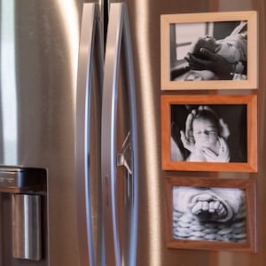 May include: Three black and white photos of a baby are displayed in wooden frames on a stainless steel refrigerator. The frames are different sizes and have different colored wood.