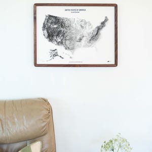 May include: A framed elevation map of the United States of America. The map is in grayscale and set against a white background. The frame is made of dark wood. The text "UNITED STATES OF AMERICA ELEVATION MAP" is at the top.