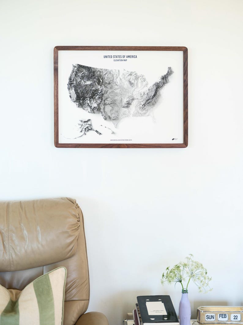 May include: A framed elevation map of the United States of America. The map is in grayscale and set against a white background. The frame is made of dark wood. The text "UNITED STATES OF AMERICA ELEVATION MAP" is at the top.