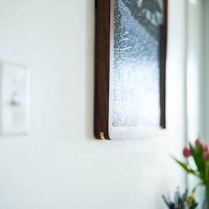 May include: A framed picture with a dark wood frame hangs on a white wall. The artwork shows a landscape with silhouetted figures against a yellow and blue backdrop. The frame has a gold accent.