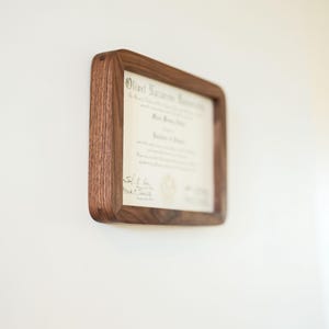 May include: A certificate displayed in a wooden frame, mounted on a white wall. The frame is a warm brown colour, square with rounded corners. The document inside is a certificate with text.