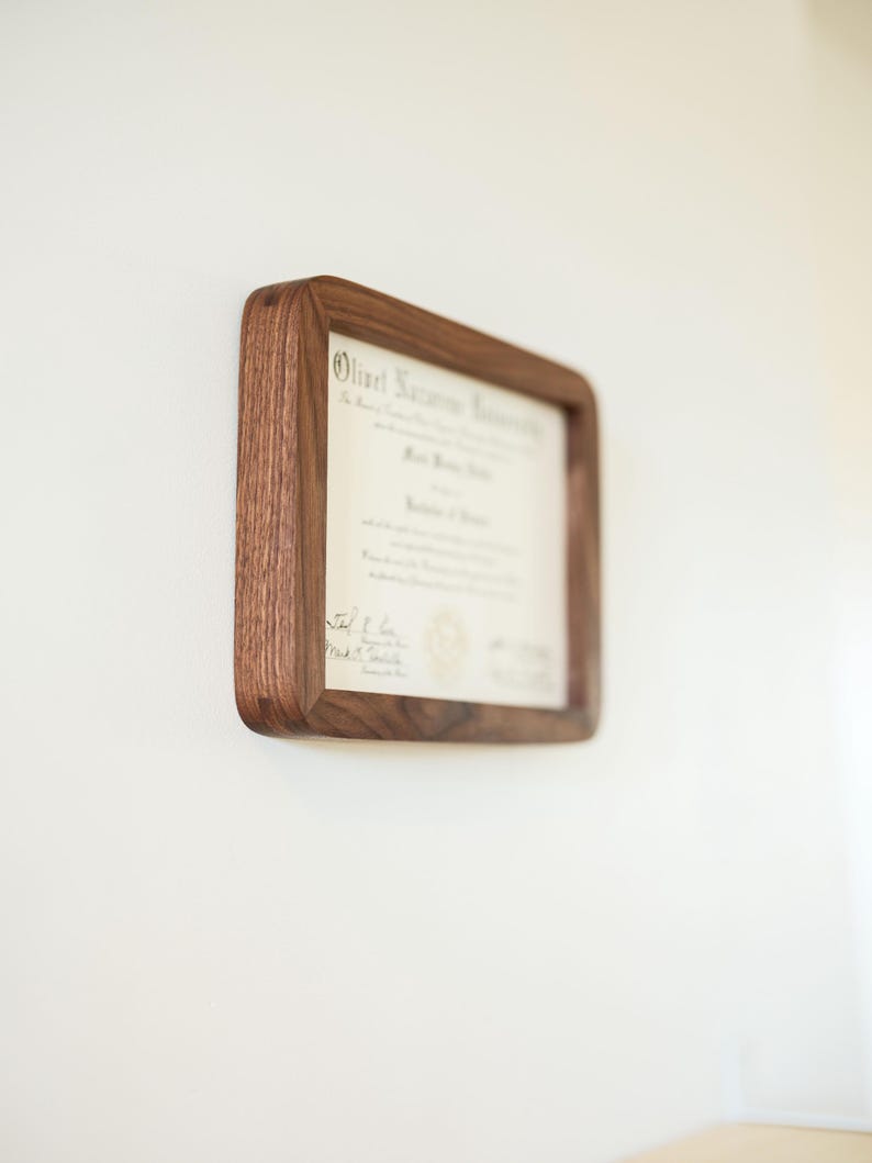 May include: A certificate displayed in a wooden frame, mounted on a white wall. The frame is a warm brown colour, square with rounded corners. The document inside is a certificate with text.
