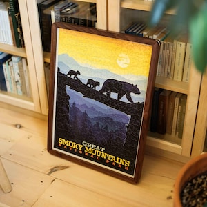 May include: A framed jigsaw puzzle depicting the Great Smoky Mountains National Park. The image shows three bears on a ridge with a sunset. The frame is made of dark wood. The puzzle is likely 50 cm x 70 cm.