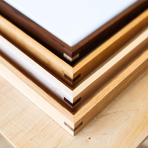 May include: Close-up of three wooden picture frames stacked on top of each other. The frames are different shades of brown and tan, with a white surface visible in the top frame. The frames are on a light wood surface.