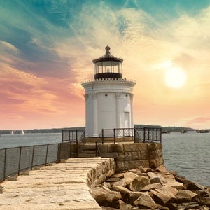 May include: A white lighthouse with a black roof and a black lantern on a rocky shore. The lighthouse is surrounded by a black metal fence. The sky is a beautiful blue and orange.