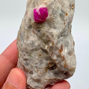 May include: A raw mineral specimen featuring a prominent, vibrant magenta-coloured crystal embedded within a textured, light grey and white matrix. The stone surface displays scattered small brown inclusions and crystalline facets throughout its structure.