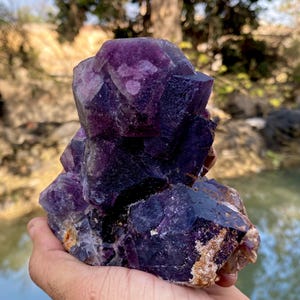 Könnte beinhalten: Ein großer, roher Amethyst-Kristallcluster mit tiefvioletten und violetten Farbtönen. Die Kristallformation weist mehrere Spitzen und Facetten auf, die ihren natürlichen, unpolierten Zustand zeigen. Die Basis hat eine hellbraune Farbe.
