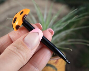 Può includere: Un'annaffiatoio in vetro nero con una parte superiore a forma di fungo giallo e nero.