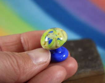 Può includere: Un piccolo fungo di vetro con un motivo a spirale giallo e blu sulla parte superiore e una base blu solida.