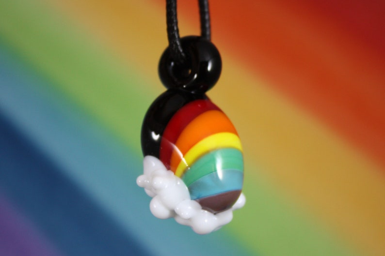 May include: A rainbow glass pendant with a black cord and a white cloud charm. The pendant features a rainbow with red, orange, yellow, green, blue, and purple stripes.